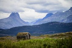 an old shack sits in the middle of a grassy field with mountains in the background