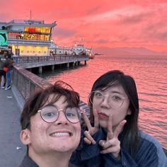 two people standing next to each other near the water at sunset with a pier in the background