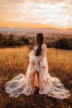 a woman in a long dress sitting on the ground with her legs crossed looking at the sunset