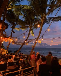 people are sitting at tables under palm trees on the beach with lights strung over them