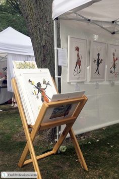 an easel with paintings on it in front of a white tent and some trees