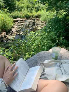 a woman reading a book while sitting next to a stream in the woods on a blanket