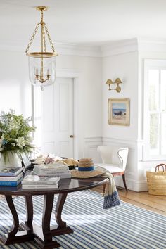 a dining room table with books on it in front of two windows and a chandelier hanging from the ceiling