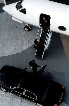 an aerial view of a black car parked in front of a white plane with people standing around it