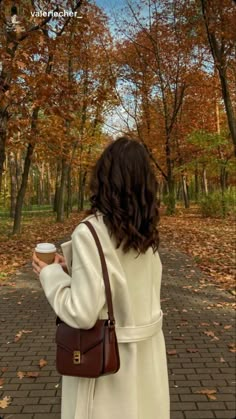 a woman in a white coat is holding a coffee cup and looking at the trees