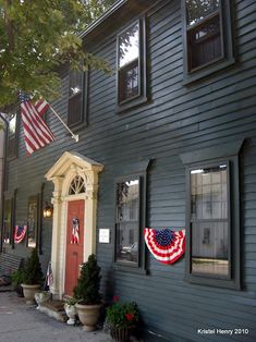 an american flag hanging on the side of a gray house with three windows and two flags