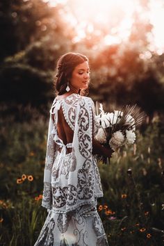 a woman in a white dress is holding flowers and looking at the sun behind her