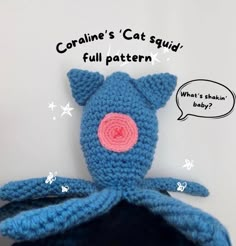 a blue crocheted stuffed animal with a pink nose and tail sitting in front of a white wall