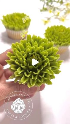 a hand holding a green flower in front of three cupcakes on a table