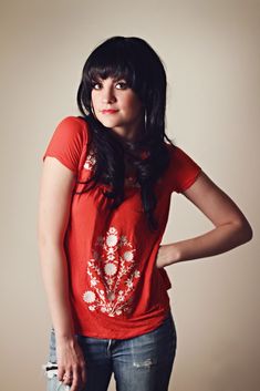 a woman with long hair wearing a red t - shirt and jeans standing in front of a wall