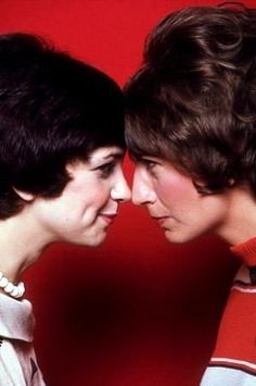 two women standing next to each other in front of a red wall and one has her head on another woman's shoulder