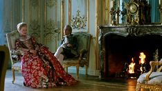 a man and woman sitting on chairs in a room with ornate wallpaper, fireplace and chandelier