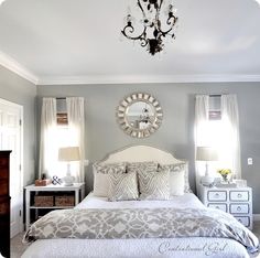 a white bed sitting under a chandelier next to two windows in a bedroom