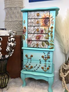 a blue dresser with flowers painted on the front and drawers, next to some dried plants