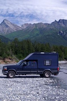 a truck is parked on the side of a river with mountains in the back ground