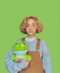 a woman in overalls holding a green stuffed frog on top of her chest and looking at the camera