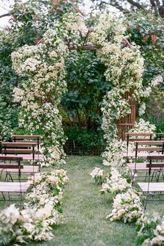 an outdoor ceremony set up with wooden chairs and white flowers on the aisle, surrounded by greenery