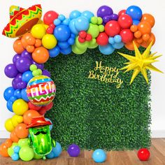 a birthday arch with balloons and confetti in front of an artificial hedge wall