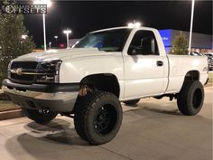 a white truck is parked in a parking lot at night with its lights on and it's big tires