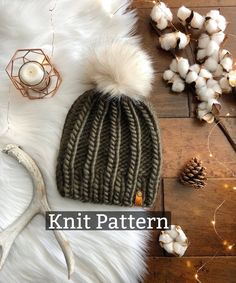 a knit beanie hat with white fur pom - poms sits on top of a wooden floor