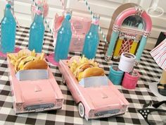 two pink cars are sitting on a checkered tablecloth with sodas and hotdogs