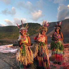 three women in grass skirts and headdresses stand on rocks near the ocean