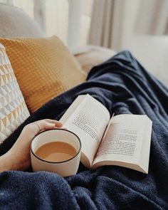 a person holding a cup of coffee and reading a book on a bed with pillows