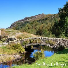 an old stone bridge over a small stream in the middle of a field with mountains behind it