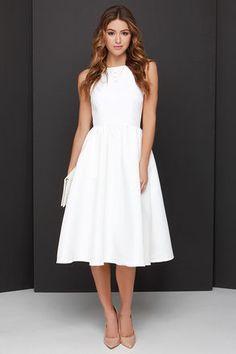 a woman wearing a white dress and heels standing in front of a black wall with her hands on her hips