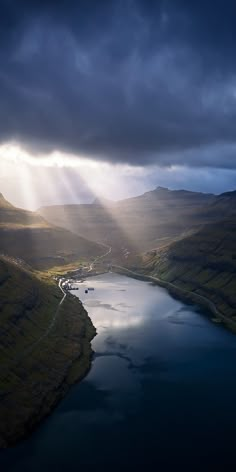 an aerial view of a lake surrounded by hills and trees under a dark sky with sun rays coming through the clouds