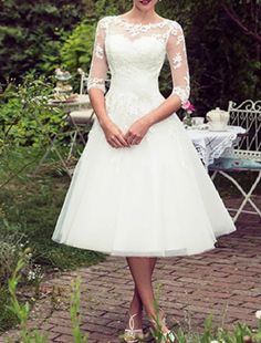 a woman in a white dress standing on a brick walkway with flowers and greenery behind her