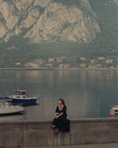 a woman sitting on the edge of a wall next to water with boats in the background