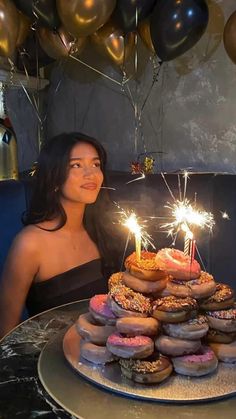 a woman sitting in front of a cake with lit candles on it and lots of doughnuts