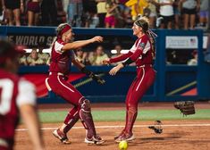 two women in red uniforms playing softball on a baseball field with fans watching from the stands