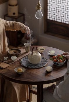 a woman pouring tea into a white teapot on top of a wooden dining table