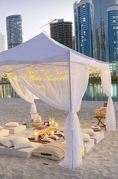 a white canopy bed sitting on top of a sandy beach next to tall city buildings