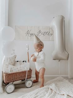 a baby in a birthday hat standing next to a wagon with balloons and presents on it