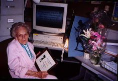 an older woman sitting in front of a computer desk with a monitor and keyboard on it