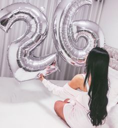 a woman sitting on top of a bed holding two silver balloons in the shape of numbers