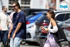 a man and woman walking down the street in front of parked cars, talking to each other