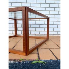 a glass and wood box sitting on the ground next to a brick wall with a small green plant growing in it