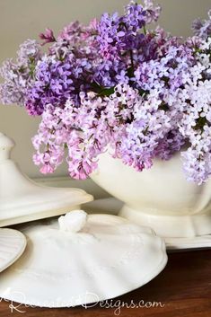 purple flowers are in a white vase on a wooden table next to seashells