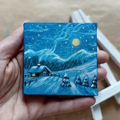 a hand holding up a small painting of a cabin in the snow with stars and moon