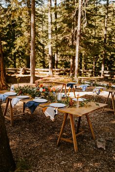 an outdoor table set up in the woods with flowers and place settings laid out on it