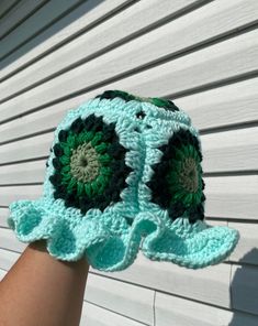 a crocheted green and black hat is held up against a white building wall