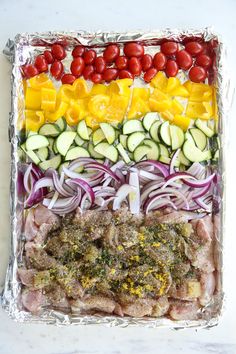 an assortment of vegetables and meat on tin foil in a rainbow - colored tray with red onions, cucumbers, yellow peppers, tomatoes, zucchini, and other veggies