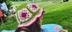 a woman sitting in the grass wearing a green and pink crocheted hat with flowers on it