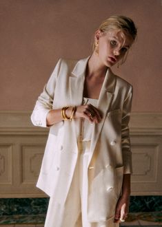 a woman standing in front of a wall wearing a white suit and gold bracelets