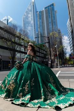 a woman in a green and gold dress standing on the sidewalk with tall buildings behind her