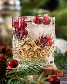 a glass filled with ice and cherries sitting on top of a wooden table next to pine cones